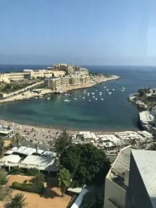 Aerial view of St. George’s Bay in Malta with a sandy beach, sun umbrellas, swimmers, boats, and hotels along the coastline.