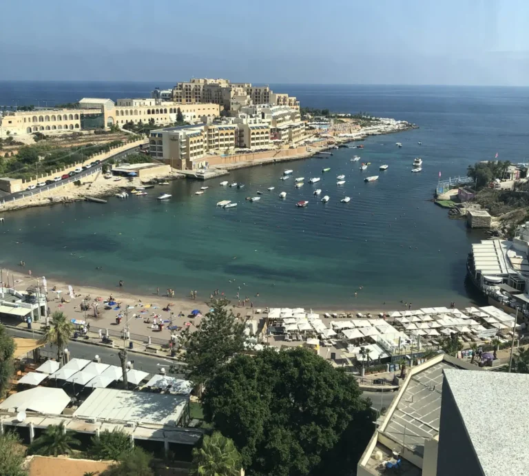 Aerial view of St. George’s Bay in Malta with a sandy beach, sun umbrellas, swimmers, boats, and hotels along the coastline.
