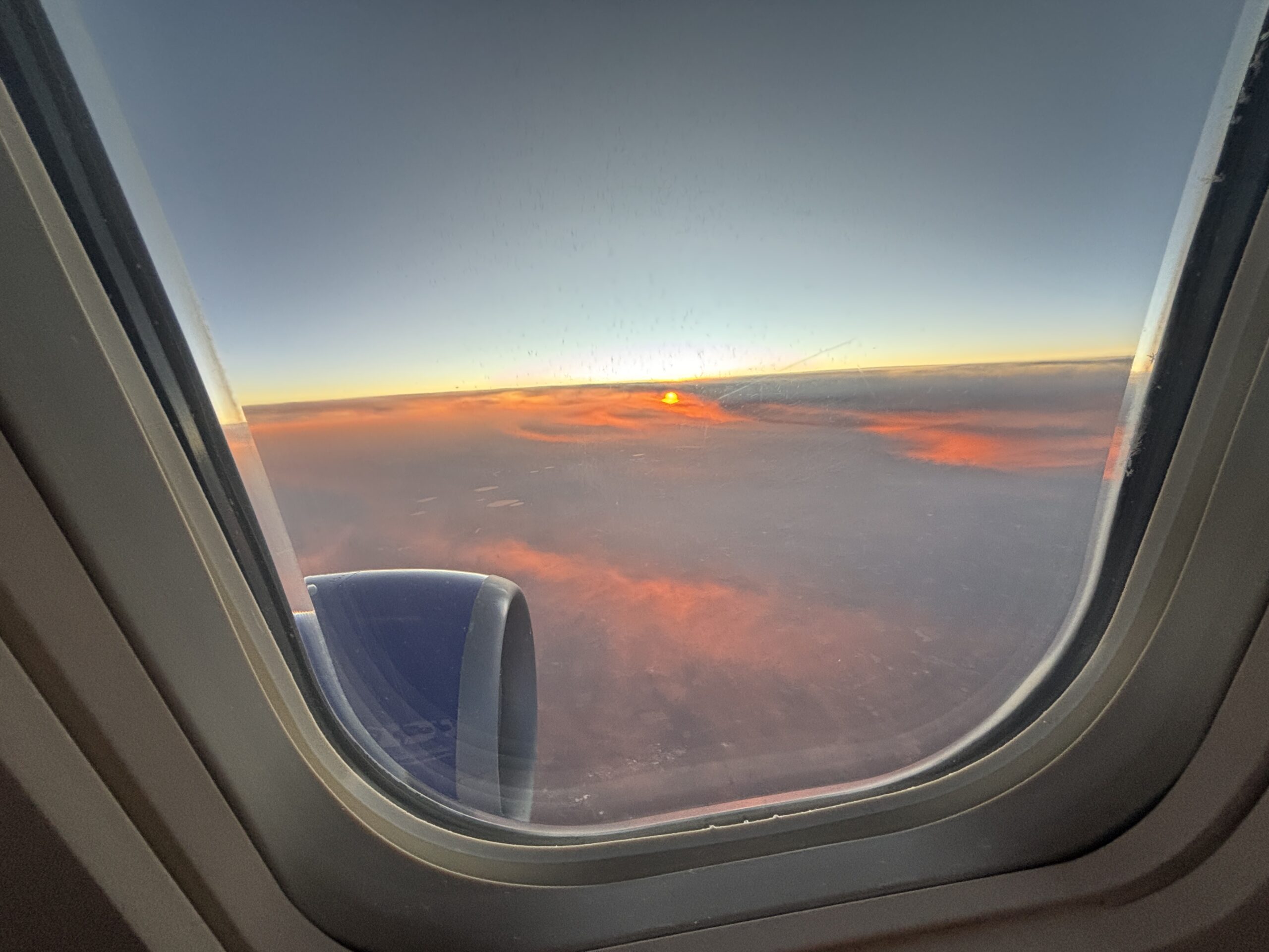 Sunset view from airplane window showing orange and pink clouds above the horizon with engine visible on the left side.