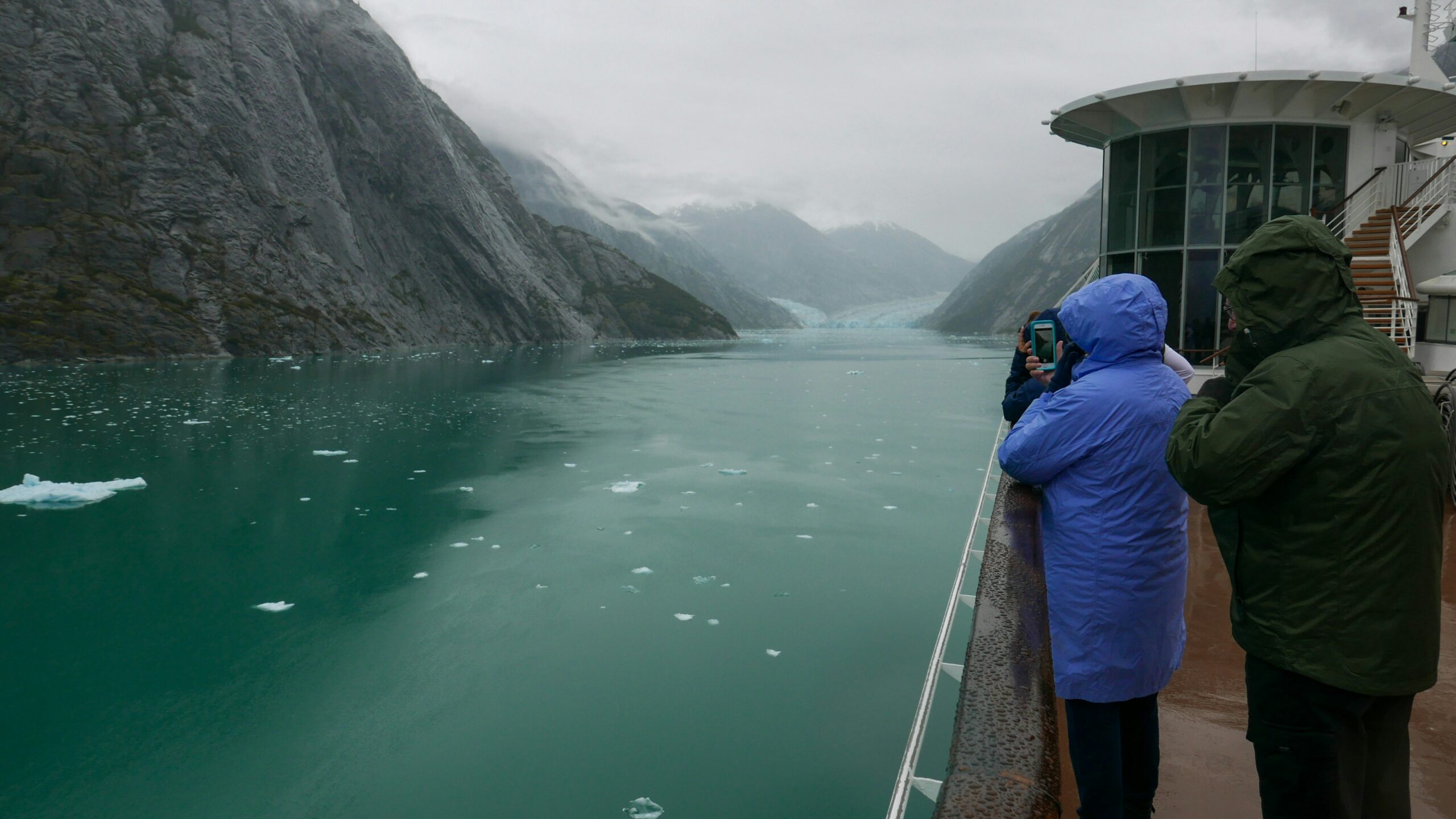 Cruise passengers in rain jackets watching a glacier at the end of a narrow Alaskan fjord