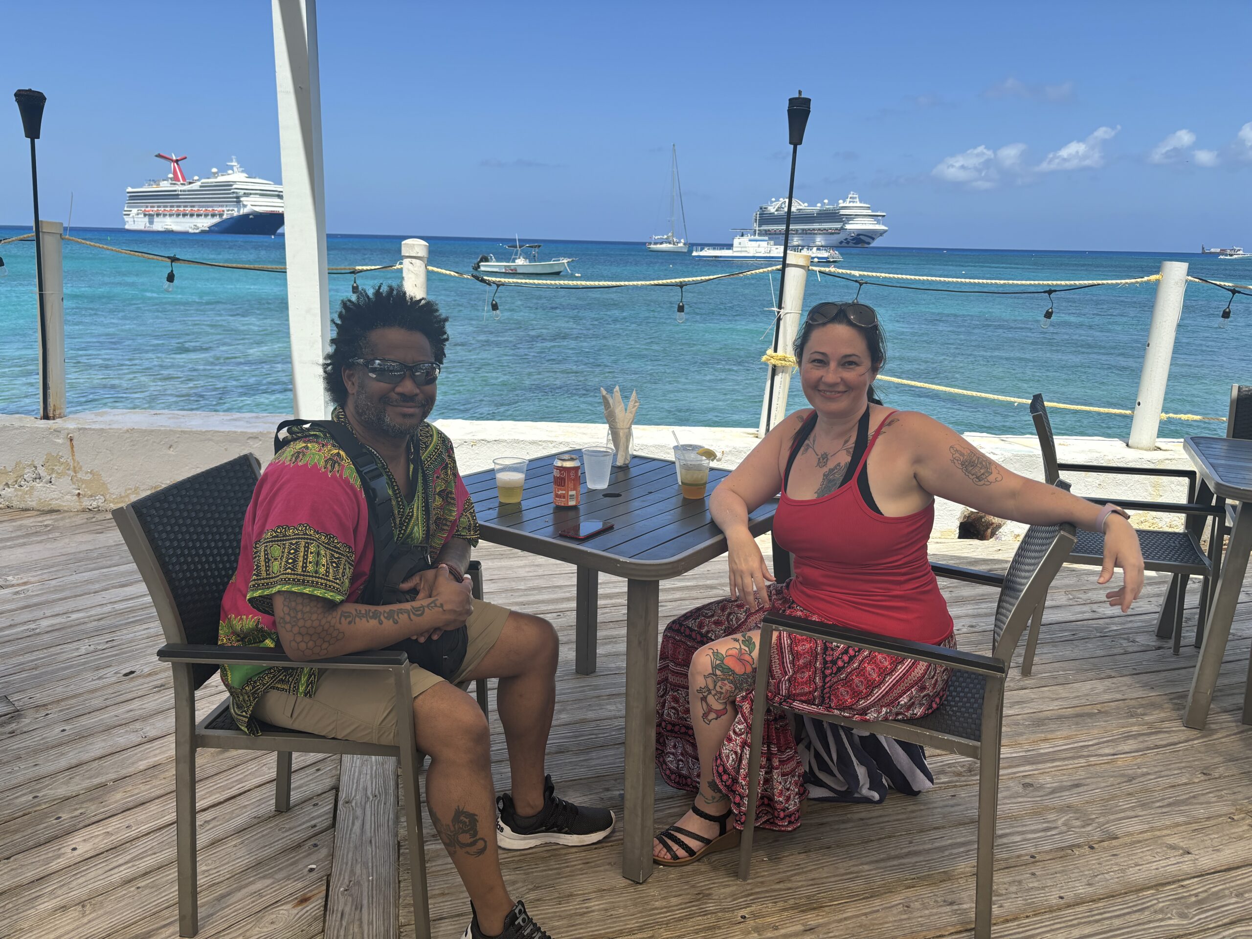 Couple at a waterfront table in Grand Cayman with cruise ships anchored offshore in turquoise water